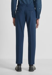 Pantaloni blu navy con una vestibilità su misura. Presentano due tasche posteriori con chiusura a bottone e una texture liscia. Indossati con scarpe nere.