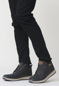 Melni potītes ar gludu apdari, šņorējamu dizainu, teksturētu mēli un gaišu zoli, valkātas kopā ar melnām biksēm.