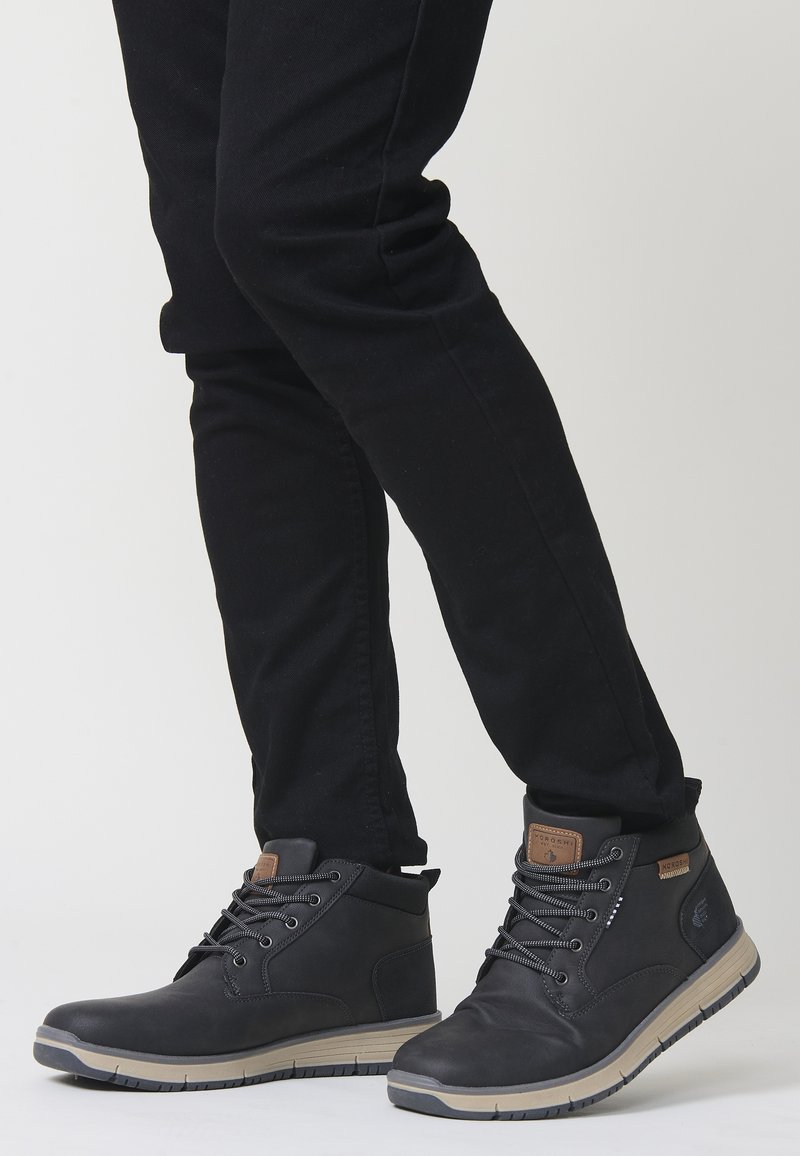 Melni potītes ar gludu apdari, šņorējamu dizainu, teksturētu mēli un gaišu zoli, valkātas kopā ar melnām biksēm.