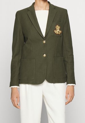 Vrouw draagt een olijfgroene blazer met gouden knopen en een geborduurd wapenschild, gecombineerd met witte hooggetailleerde broek en een wit topje.