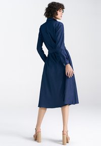 Sukienka dżinsowa w ciemnoniebieskim kolorze, z długimi rękawami, przewężona w talii paskiem, kołnierz, długość do połowy łydki, z falującą spódnicą i guzikami przy mankietach.