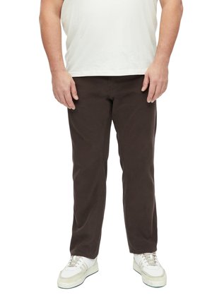 Mann trägt dunkelbraune Hose, weiße Sneakers und ein weißes Hemd und steht vor weißem Hintergrund.