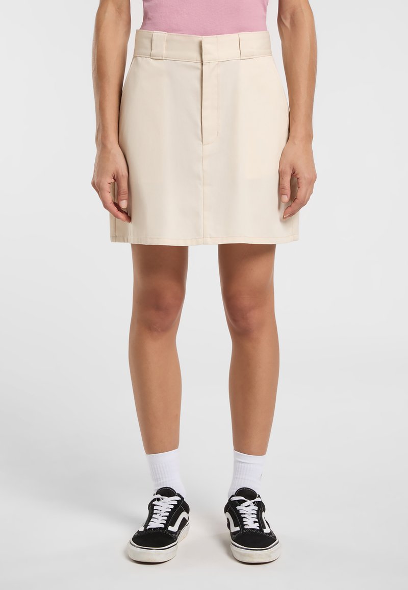 Beige bomullsblandad kjol med rak skärning, framficka, platt midja och en veck längs framsidan, kombinerad med svarta sneakers och vita strumpor.