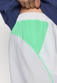 Λευκά αθλητικά σορτς με πράσινη  μέντα διχτυωτή επένδυση, ελαστική μέση και επίπεδα ραφές. Το υλικό φαίνεται ελαφρύ και αεριζόμενο.