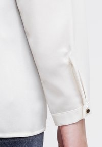 Poignet de chemise blanche à manches longues avec une petite fermeture par bouton, portée par-dessus un jean en denim bleu, montrant une partie du bras et de la taille.
