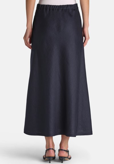 Femeie purtând o fustă lungă evazată bleumarin închis, cu talie cu elastic și sandale negre cu toc mic și deschis la vârf, văzută din spate.