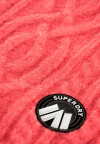 Tejido de punto rosa con una textura suave y esponjosa. Presenta un parche circular negro con "SUPERDRY" y un diseño geométrico en blanco.