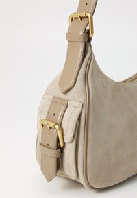 Bežinis zomšinis rankinės su mažu sagties kišenėle ir reguliuojamu odiniu dirželiu, papuoštu auksine metaline sagtimi.