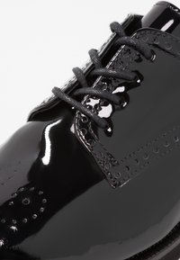 Czarne lakierowane buty skórzane z dekoracyjnymi perforacjami, teksturowanymi sznurówkami i błyszczącym wykończeniem. Posiadają okrągły nosek i kontrastowe przeszycia.