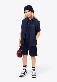 Lapsi, joka on pukeutunut laivansinisellä pipoa, ilmanhihaisessa toppaliivissä, paidassa ja shortseissa, pitää kädessään pientä marjakuosisia käsilaukkua ja seisoo yksi käsi taskussa.