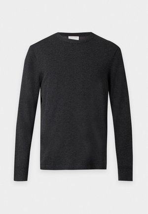 Homme debout face à l’avant portant une chemise à manches longues gris foncé, un pantalon noir large et des chaussures noires sur un fond blanc uni.