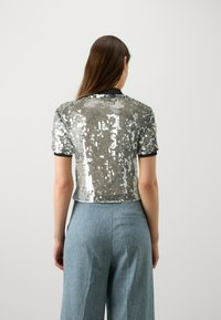 Top corto a maniche corte in paillettes argento con colletto nero e design corto, abbinato a pantaloni a gamba larga di un blu chiaro. Texture liscia con riflessi.