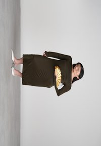 Γυναίκα με φόρεμα σε ελιά πράσινο με μακριά μανίκια, κρατώντας ένα χρυσό αναδιπλούμενο ανεμιστήρα, φορώντας λευκές ψηλές μπαλαρίνες, στέκεται μπροστά σε έναν ουδέτερο φόντο.