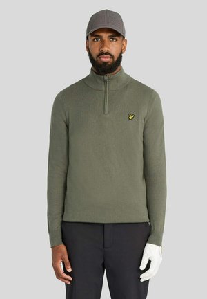 Homme portant un pull zippé vert olive, une casquette grise perforée, un pantalon noir et un gant de golf blanc à la main droite sur un fond gris uni.
