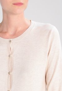 Ljust beige cardigan i mjukt stickat material, med rund halsringning, fem knappstängning och subtil ribbad textur.