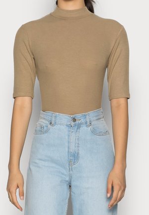 Nainen, jolla on päällään istuva beige, ribbattu puolikaulusneule kyynärpään pituisilla hihoilla yhdistettynä korkeavyötäröisiin vaaleansinisiin farkkuihin.