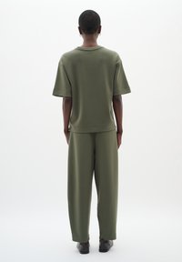 Osoba s krátkými vlasy a kroužkovými náušnicemi, oblečená v volném olivově zeleném topu s krátkým rukávem a odpovídajících kalhotách, stojící na pozadí v jednoduché bílé barvě.