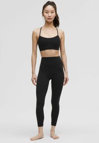 Schwarzer Sport-BH mit dünnen Trägern, kombiniert mit hochsitzenden schwarzen Leggings. Beides aus glattem, dehnbarem Material gefertigt.