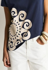 Granatowy bawełniany T-shirt z dużym beżowym wzorem w kształcie spirali po jednej stronie, uzupełniony białymi spodniami z denimu oraz wieloma bransoletkami.