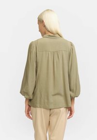 Blusa verde oliva con maniche a sbuffo e design a pieghe sul retro, realizzata in tessuto leggero, abbinata a pantaloni beige.