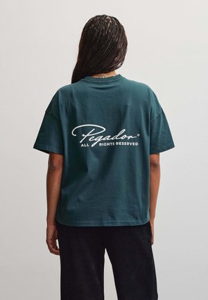 Person med fletninger iført en mørkegrøn T-shirt med teksten "Pegador All Rights Reserved" på ryggen, kombineret med sorte bukser.