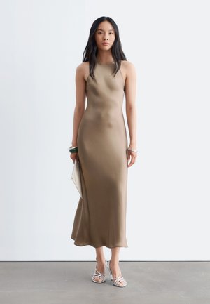 Femme aux longs cheveux foncés portant une robe taupe sans manches longue jusqu'aux chevilles, tenant une pochette beige et portant des talons blancs à brides.