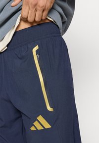 Teget sportske kratke hlače sa žutom bočnom prugom i džepom na patentni zatvarač. Imaju elastični pojas i Adidas logo u zlatnoj boji. Glatke teksture.