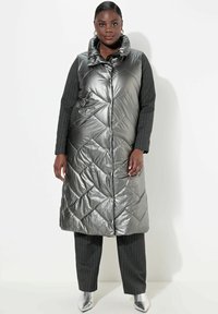 Metallisch graune gepolsterte Weste mit hohem Kragen, ausgestattet mit einem Rautenmuster, Reißverschluss und Seitentaschen. Kombiniert mit Nadelstreifenhosen.