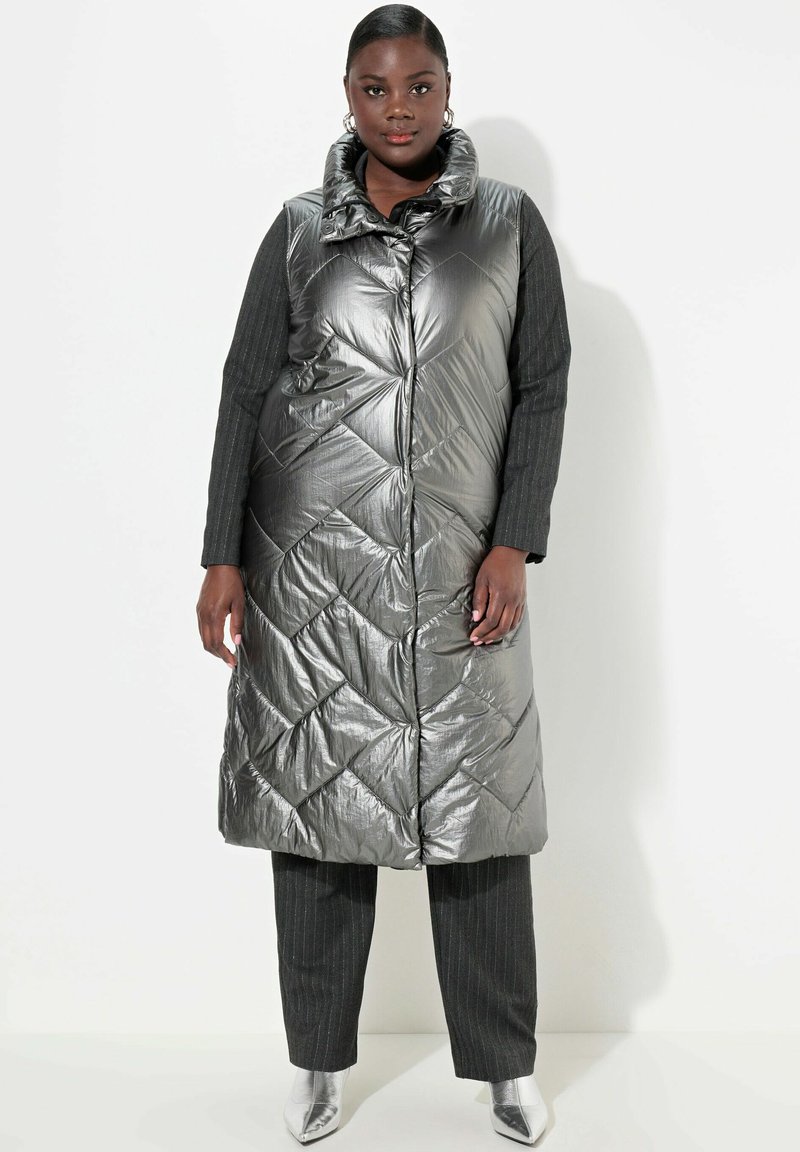 Metallisch graune gepolsterte Weste mit hohem Kragen, ausgestattet mit einem Rautenmuster, Reißverschluss und Seitentaschen. Kombiniert mit Nadelstreifenhosen.