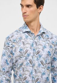 Man in een lichtblauw bloemetjes overhemd met blauwe en grijze bladeren en bloemen, kijkend naar links tegen een witte achtergrond.