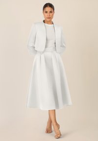 Biały trzyczęściowy garnitur składający się z dopasowanej, krótkiej marynarki, bluzki z krótkim rękawem i zwiewnej spódnicy. Gładka tekstura z plisami. Przezroczyste sandały.