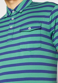 Zeleno-modré pruhované polo tričko s knoflíkovým límcem, krátkými rukávy a kapsou na prsou. Obsahuje světle modré knoflíky.