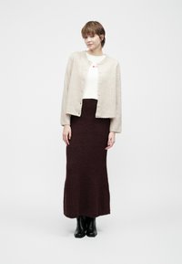 Vrouw in een beige knoopcardigan over een witte top, met een donkerpaarse lange rok en zwarte laarzen, staande tegen een eenvoudige witte achtergrond.