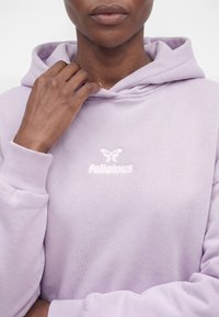 Personne portant un sweat à capuche violet clair avec le texte « felicious » et un logo papillon, tenant le sweat près du cou avec une main.