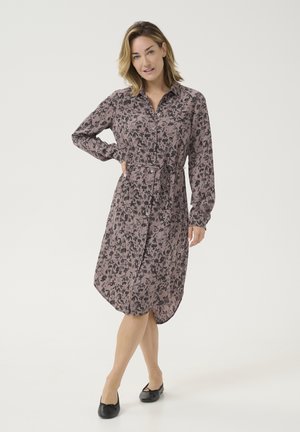 Abito a camicia in morbido rosa e nero con stampa floreale, caratterizzato da una vita con fiocco, maniche lunghe e orlo asimmetrico. Scarpe flat nere.