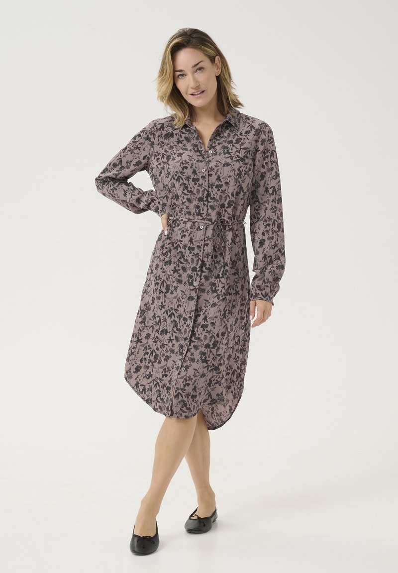 Robe à boutons imprimée florale en rose doux et noir, avec une taille à nouer, des manches longues et un ourlet asymétrique. Chaussures plates noires.