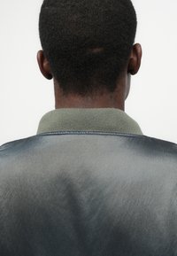 Jaqueta bomber em um tecido liso, azul-escuro e cinza. Apresenta um colarinho canelado verde e costuras visíveis ao longo da linha do pescoço.