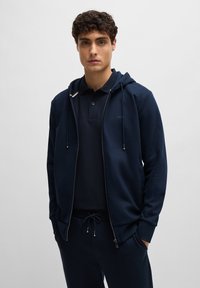 Sweatshirt azul-marinho com fecho éclair e capuz ajustável, combinado com uma t-shirt polo a condizer; apresenta um corte slim e um toque suave.
