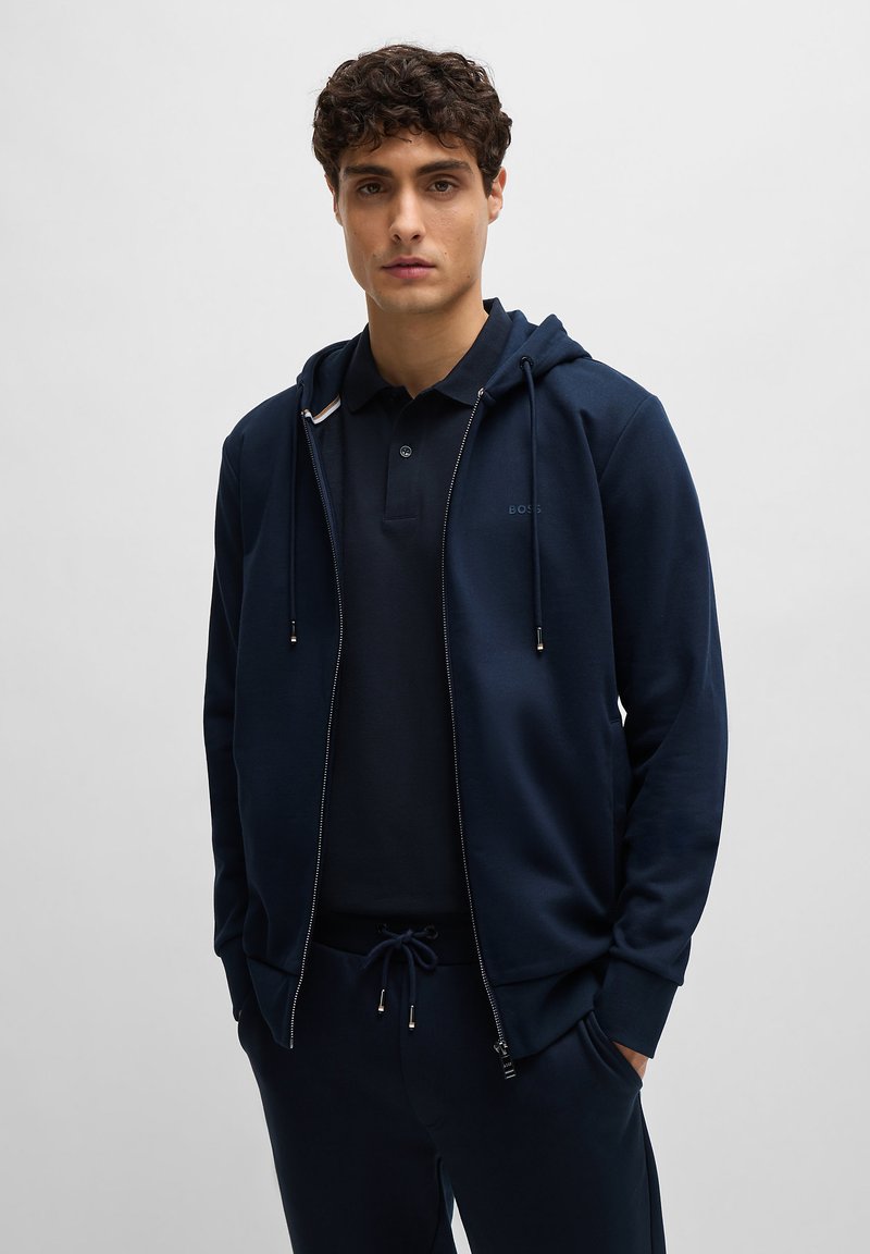 Sweatshirt azul-marinho com fecho éclair e capuz ajustável, combinado com uma t-shirt polo a condizer; apresenta um corte slim e um toque suave.