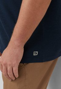 Marineblauwe shirt met structuur en korte mouwen, met een klein label aan de zoom, gecombineerd met lichtbruine broek.