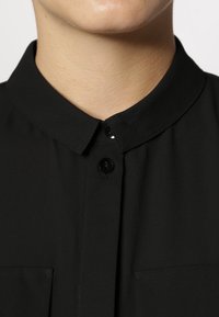 Närbild av en persons underansikte och nacke som bär en svart skjorta med krage och två synliga svarta knappar.