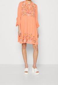 Korallefarbenes Kleid mit floralem Stickerei, Rüscheärmeln und gebundenem Ausschnitt. Kombiniert mit weißen, offenen Plateau-Schuhen. Weicher, fließender Stoff.