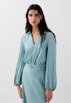 Blusa azzurra chiara, strutturata, con scollatura a V e maniche lunghe e voluminose. Presenta una vita arricciata per un look aderente.