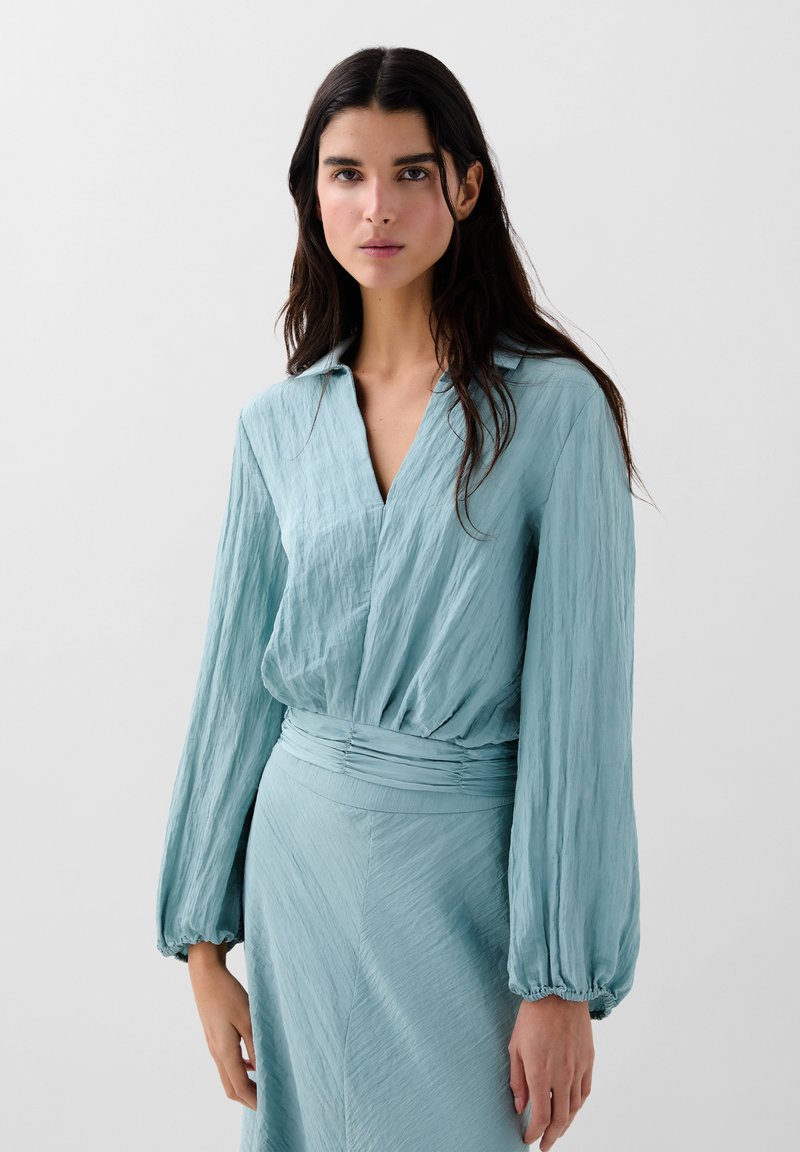 Lyseblå, tekstureret bluse med V-udskæring og lange, voluminøse ærmer. Har en samlet talje for et figursyet look.