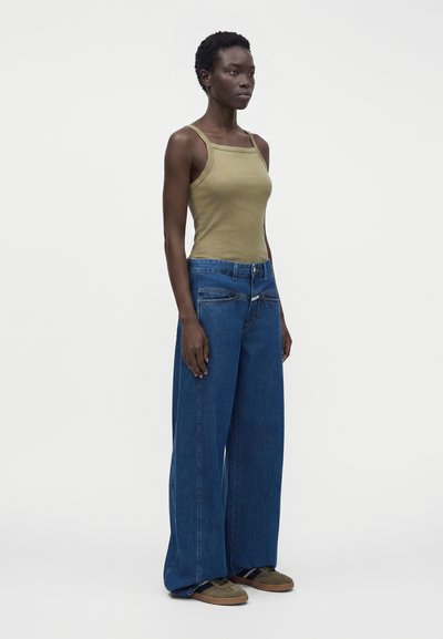 Femme debout portant un débardeur vert olive, un jean large bleu et des baskets vert olive sur un fond clair uni.