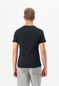Tânăr cu păr scurt și creț, purtând un tricou negru simplu și blugi gri deschis, întors cu spatele, pe un fond alb.