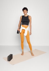 Top curto preto, leggings laranja e sweatshirt branca amarrada na cintura. Descalça, segurando uma bolsa preta. Tapete de yoga de cor clara por perto.