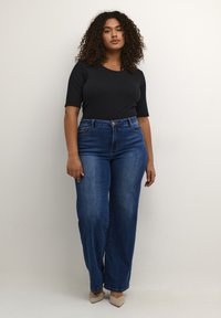 Top negro ajustado con mangas cortas, combinado con vaqueros rectos de tiro alto en azul. La modelo lleva tacones beige, con un fondo neutro.