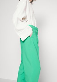 Persoon in een losse, witte, lange mouwen shirt dat is gestopt in felgroene hoge taillebroek met handen in de zakken, tegen een effen achtergrond.