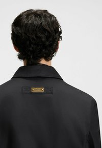 Black coat featuring a wide collar, textured fabric, and a gold-tone "HUGO" logo tag on the back near a decorative stitching detail.